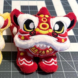 Puede incluir: Un juguete de felpa de danza del león rojo y blanco con detalles en negro, amarillo y dorado. El juguete tiene ojos grandes, una boca ancha y bordados detallados. El juguete de danza del león es un símbolo cultural de buena suerte y prosperidad.