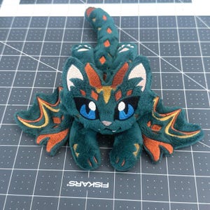 May include: A teal plush creature with large blue eyes, white accents, and orange and gold details. The plush has a long tail and wing-like appendages. The creature is lying on a gray cutting mat.