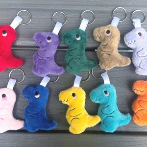 May include: A collection of ten plush dinosaur keychains in various colors, including red, purple, green, tan, light purple, pink, blue, yellow, teal, and orange. Each keychain features a silver ring and a white fabric loop.