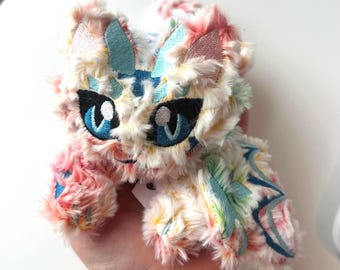 Op de afbeelding: Een pluche kat met een pluizig, veelkleurig uiterlijk. De kat heeft geborduurde details, waaronder grote blauwe ogen, zwarte accenten en blauwe en roze accenten op de oren en vleugels. Het speelgoed wordt in een hand gehouden.