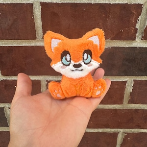 May include: A plush orange fox toy with white accents on the face and ears, and light blue eyes. The fox is sitting with its paws forward. The toy is held in a hand against a brick background.