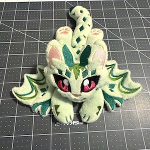 May include: A plush, light green cat-like creature with large pink eyes and a patterned design. The creature has a long tail and decorative wings. The plush toy is made of soft material and has embroidered details.