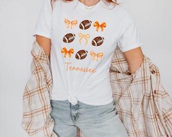 May include: White t-shirt with orange and brown bows and footballs. The text "Tennessee" is printed on the shirt.