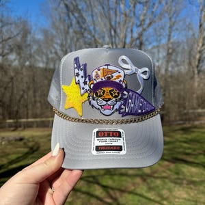 May include: Gray trucker hat with a purple and yellow tiger graphic, a gold chain, and a white bow. The hat has a label that reads "OTTO World Famous Foam Front OSFM Trucker Durable Snap Closure".