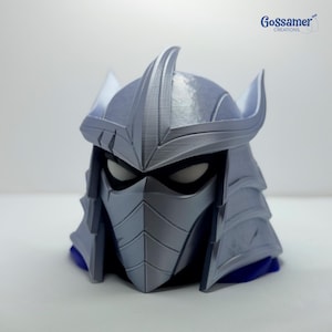 May include: A silver-colored, 3D-printed helmet featuring a pointed crest, angular face mask, and dark eye openings. The design includes layered plating details and a sharp, spiked silhouette. The text Gossamer CREATIONS appears in the top right corner.