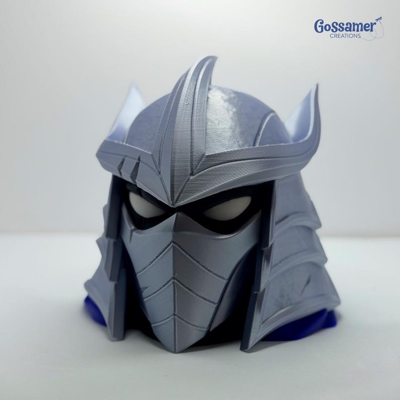 May include: A silver-colored, 3D-printed helmet featuring a pointed crest, angular face mask, and dark eye openings. The design includes layered plating details and a sharp, spiked silhouette. The text Gossamer CREATIONS appears in the top right corner.