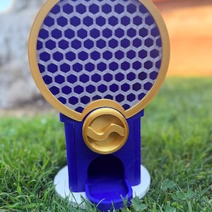 May include: A blue and gold gumball machine featuring a circular top section with a white honeycomb pattern behind a gold frame. The base is blue with a gold circular dial in the center and a small dispensing chute at the bottom on a white stand.