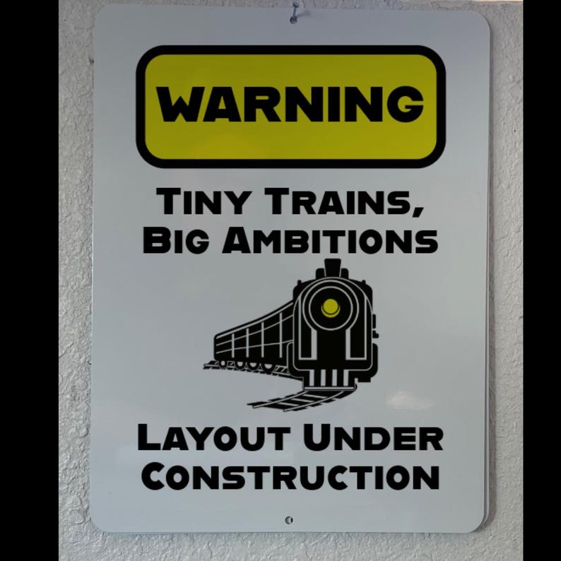 Pu&ograve; includere: Un cartello bianco con testo nero che recita "ATTENZIONE TRENI PICCOLI, GRANDI AMBIZIONI LAYOUT IN COSTRUZIONE". Un'illustrazione in bianco e nero di un treno si trova al centro del cartello.