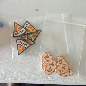 K&ouml;nnte beinhalten: Zwei durchsichtige Plastikbeutel mit kleinen Spielmarken. Ein Beutel enth&auml;lt dreieckige, pizzaf&ouml;rmige Marken mit roten Pepperoni-Kreisen und dem Text +1/+1. Der andere Beutel enth&auml;lt ovale, hellbraune Marken mit einem dunkelbraunen Grafikdesign.
