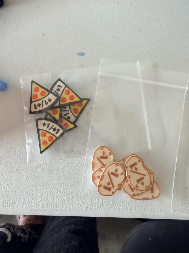 K&ouml;nnte beinhalten: Zwei durchsichtige Plastikbeutel mit kleinen Spielmarken. Ein Beutel enth&auml;lt dreieckige, pizzaf&ouml;rmige Marken mit roten Pepperoni-Kreisen und dem Text +1/+1. Der andere Beutel enth&auml;lt ovale, hellbraune Marken mit einem dunkelbraunen Grafikdesign.