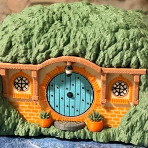 May include: A miniature dwelling featuring a bright blue circular door, orange brick-patterned walls, and two small round windows with black lattice frames. The structure is covered in textured green foliage and includes two orange planters.
