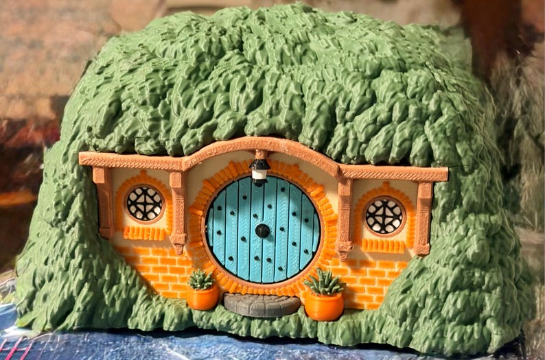 May include: A miniature dwelling featuring a bright blue circular door, orange brick-patterned walls, and two small round windows with black lattice frames. The structure is covered in textured green foliage and includes two orange planters.