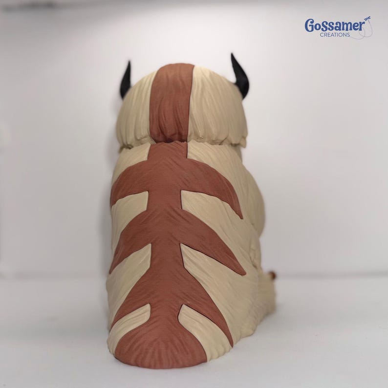 May include: A figurine of a bison-like creature, viewed from the back. The figure is primarily beige with brown markings on its back and head. It has two black horns and a tail. The figurine is a stylized representation of a character from a popular animated series.
