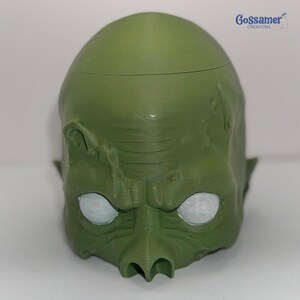 May include: A green, sculpted head with a textured surface and white eyes. The head has a prominent nose, furrowed brow, and a slight smirk. The top of the head has a seam. The logo "Gossamer Creations" is in the upper right corner.