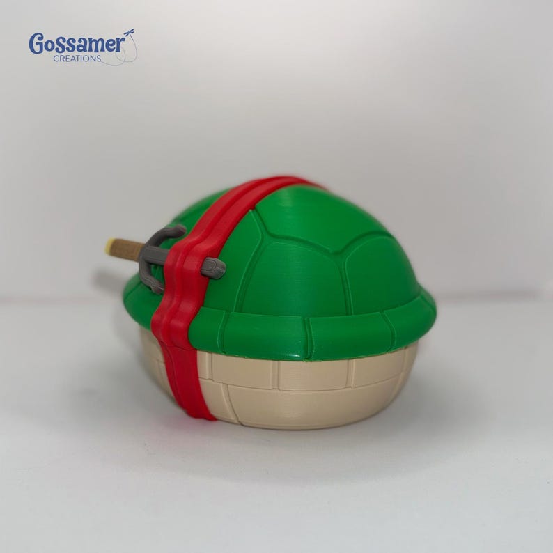 May include: A green turtle-shaped figurine with a red headband and a weapon. The figurine has a tan base and a green shell with a textured pattern. The weapon is gray and brown.