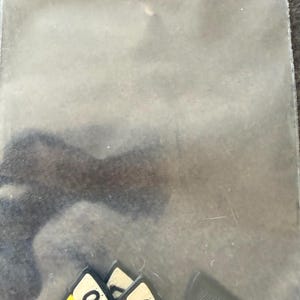 May include: A clear plastic resealable bag containing several triangular game tokens. The tokens feature a yellow and white pizza slice design with red pepperoni circles and the black text 0/+1 printed on the surface, alongside solid grey tokens.