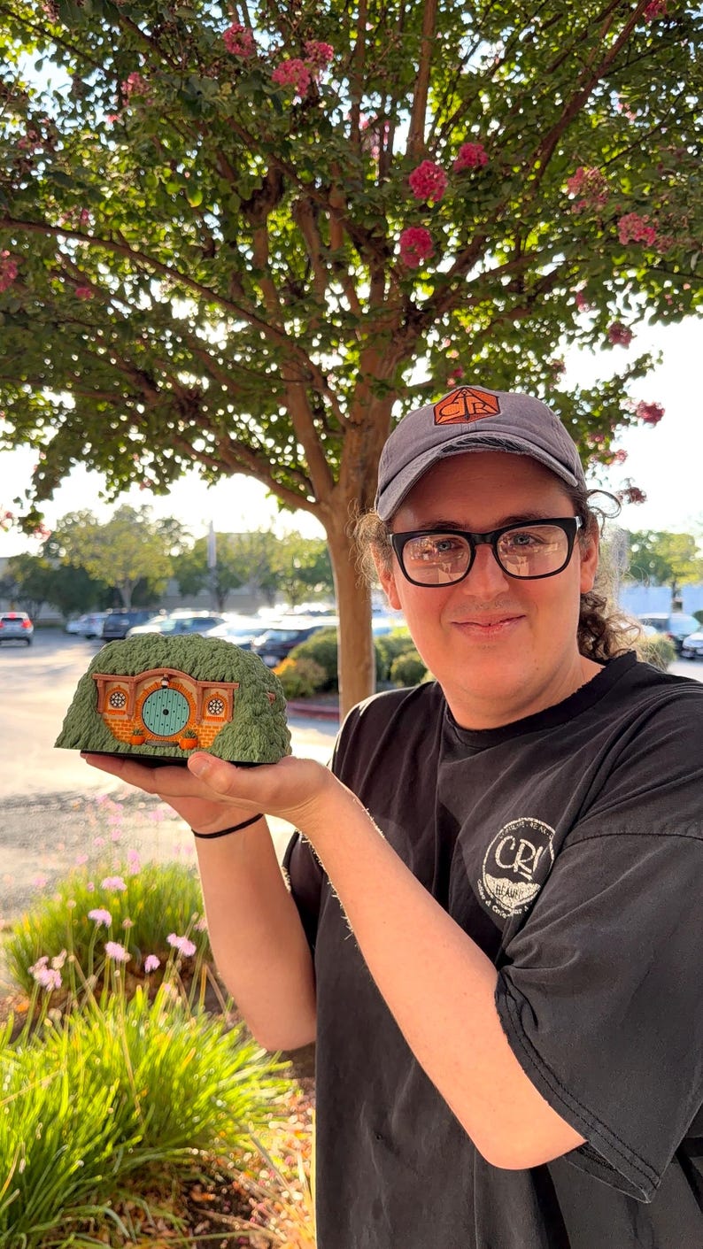 May include: A miniature hobbit-style house figurine featuring a textured green grassy roof, a round light blue front door with a central knob, and two small windows on either side of the door, all set within a brick-patterned exterior wall.
