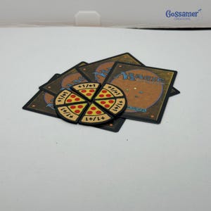 May include: A set of six pizza slice-shaped tokens with black borders, yellow crusts, and red pepperoni dots, each marked with +1/+1. These tokens are arranged in a circle on top of several Magic: The Gathering trading cards with brown backs.