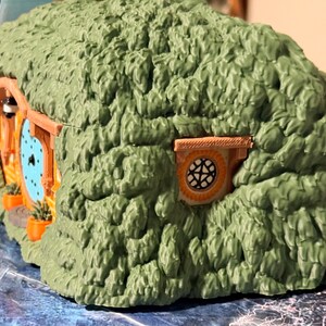 May include: A miniature hobbit-style dwelling featuring a textured green exterior resembling a grassy hill. The structure includes a round light blue door with a black handle, a small circular window with a grid pattern, and orange planters.