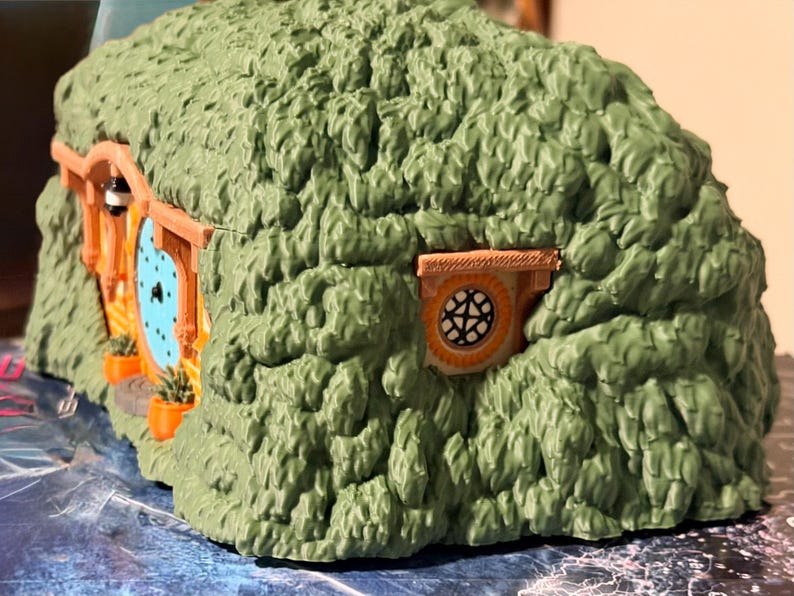 May include: A miniature hobbit-style dwelling featuring a textured green exterior resembling a grassy hill. The structure includes a round light blue door with a black handle, a small circular window with a grid pattern, and orange planters.
