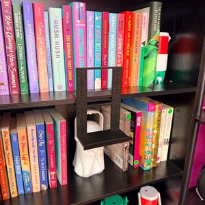 K&ouml;nnte beinhalten: Ein schwarzes, rechteckiges, 3D-gedrucktes Regal mit einem dekorativen geometrischen Muster auf der vertikalen St&uuml;tze, das auf einer wei&szlig;en, geisterf&ouml;rmigen Basis vor einem B&uuml;cherregal mit bunten B&uuml;chern und kleinen K&uuml;hlboxen steht.
