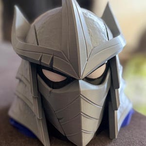 May include: A gray, 3D-printed helmet featuring a pointed crest, angular side fins, and a face mask with horizontal lines. The helmet has white eye cutouts framed by black borders, with a textured surface finish and a blue base visible at the bottom.