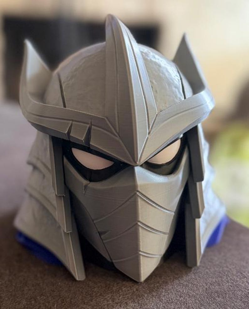 May include: A gray, 3D-printed helmet featuring a pointed crest, angular side fins, and a face mask with horizontal lines. The helmet has white eye cutouts framed by black borders, with a textured surface finish and a blue base visible at the bottom.