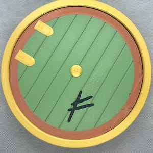 May include: A circular green door with diagonal wooden planks, a brown frame, and a bright yellow outer border. The door features two gold-colored metal hinges on the left, a central gold knob, and a black symbol resembling a stylized rune.