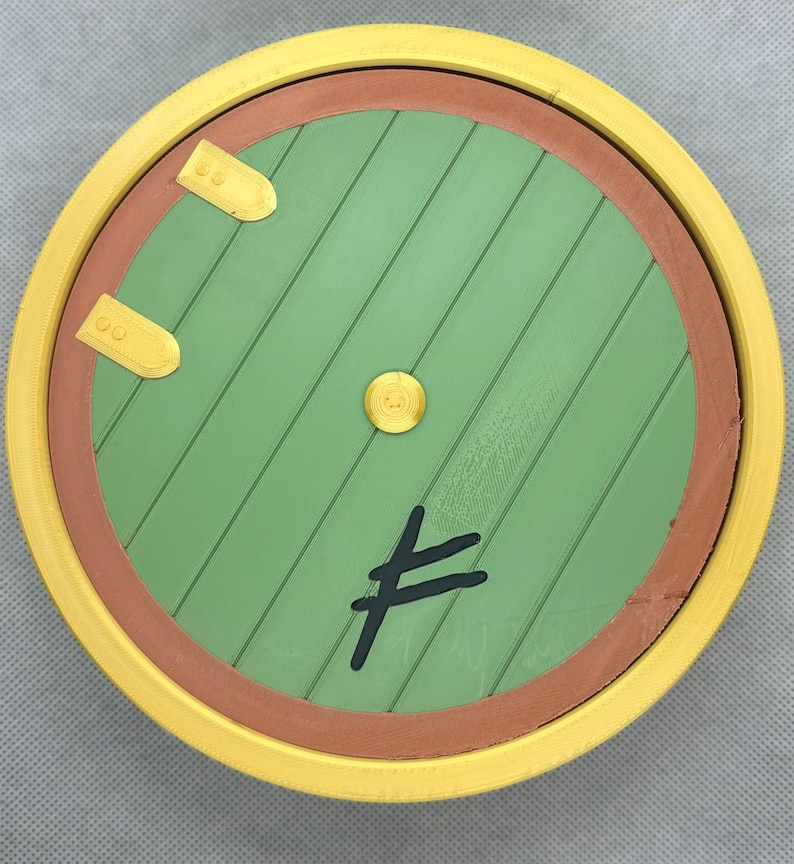 May include: A circular green door with diagonal wooden planks, a brown frame, and a bright yellow outer border. The door features two gold-colored metal hinges on the left, a central gold knob, and a black symbol resembling a stylized rune.