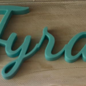 May include: A teal-colored, three-dimensional script sign displaying the name Tyra in a cursive font. The letters are connected and feature a smooth, matte finish, standing out with a bold, rounded profile against the surface beneath it.