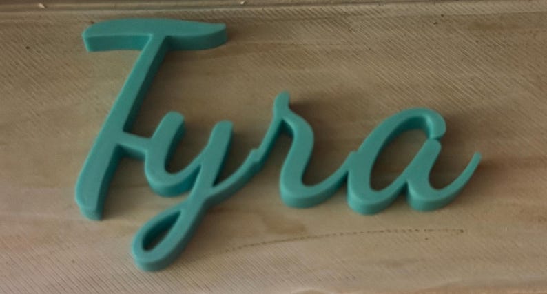 May include: A teal-colored, three-dimensional script sign displaying the name Tyra in a cursive font. The letters are connected and feature a smooth, matte finish, standing out with a bold, rounded profile against the surface beneath it.