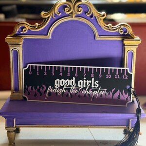 May include: A purple and gold display stand with an ornate arched top and four small feet. Resting in front is a black bookmark with a 12-inch ruler scale, purple flame graphics, and the white text "good girls finish the chapter."