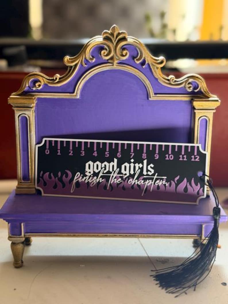 May include: A purple and gold display stand with an ornate arched top and four small feet. Resting in front is a black bookmark with a 12-inch ruler scale, purple flame graphics, and the white text "good girls finish the chapter."