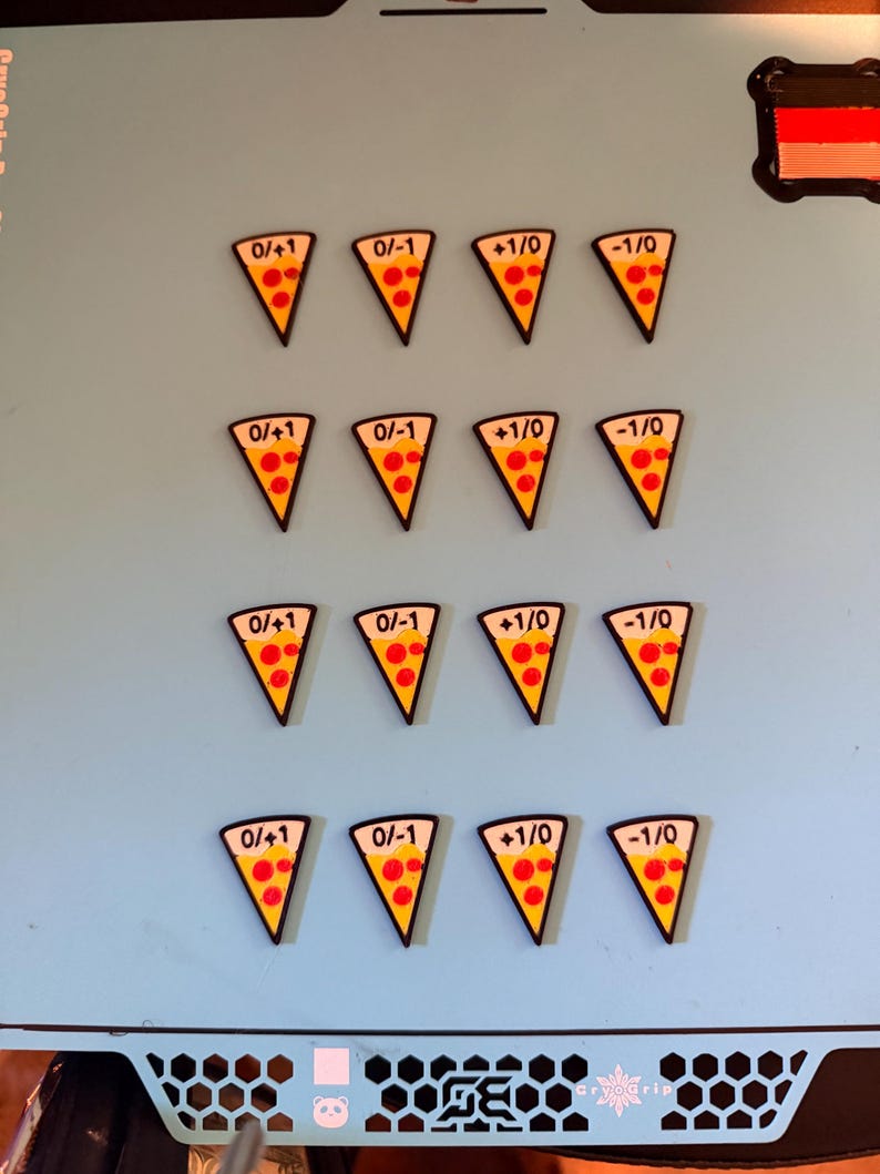 May include: Sixteen triangular pizza slice tokens arranged in four rows of four. Each token features a yellow crust, red pepperoni circles, and black text reading 0/+1, 0/-1, +1/0, or -1/0. The tokens have a black border and are set on a light blue surface.
