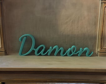 May include: A teal-colored, three-dimensional script sign featuring the name Damon in a cursive font. The letters are connected in a single piece, displaying a smooth, matte finish and a consistent thickness throughout the entire word structure.