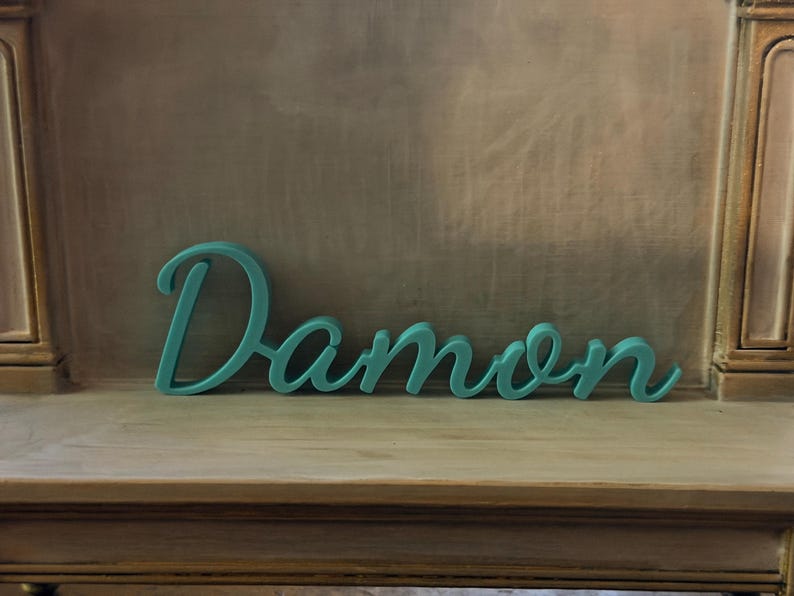 May include: A teal-colored, three-dimensional script sign featuring the name Damon in a cursive font. The letters are connected in a single piece, displaying a smooth, matte finish and a consistent thickness throughout the entire word structure.