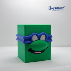 May include: A green cube-shaped decorative item featuring a blue mask and a smiling face with white eyes and a pink tongue. The item is a 3D-printed design, with the text "Gossamer Creations" in the upper right corner.