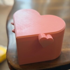 May include: A light pink heart-shaped box with a lid. The box features a small, protruding heart-shaped detail on the side near the hinge. The surface has a subtle, layered texture consistent with 3D printing, and the overall form is smooth.