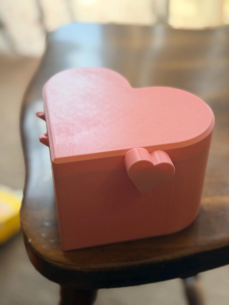 May include: A light pink heart-shaped box with a lid. The box features a small, protruding heart-shaped detail on the side near the hinge. The surface has a subtle, layered texture consistent with 3D printing, and the overall form is smooth.