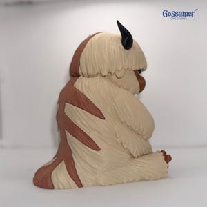 May include: A figurine of a bison-like creature with tan fur, brown markings, and black horns. The figurine is sitting down with its head turned to the side. The text "Gossamer Creations" is in the upper right corner.