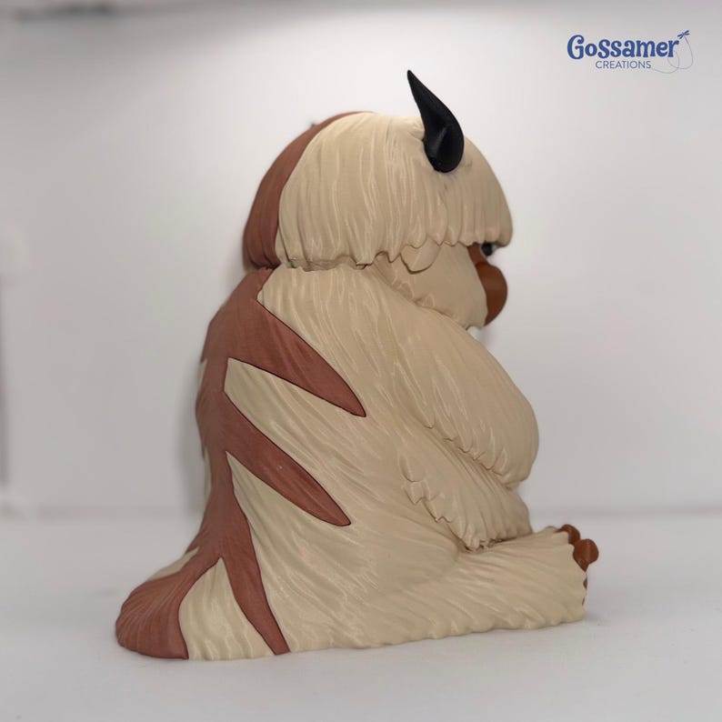 May include: A figurine of a bison-like creature with tan fur, brown markings, and black horns. The figurine is sitting down with its head turned to the side. The text "Gossamer Creations" is in the upper right corner.