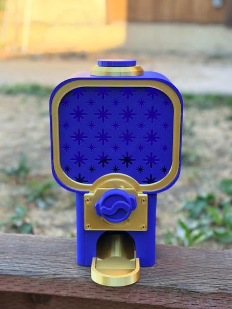 May include: A blue and gold gumball machine featuring a rectangular container with a star pattern on the front panel. The device includes a gold-coloured dispensing mechanism, a circular top lid, and a small gold tray at the base for items.