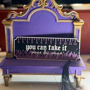 May include: A purple and gold display stand featuring a rectangular bookmark with a black and purple flame pattern. The bookmark displays a 12-inch ruler along the top edge and the text "you can take it page by page" with a black tassel.