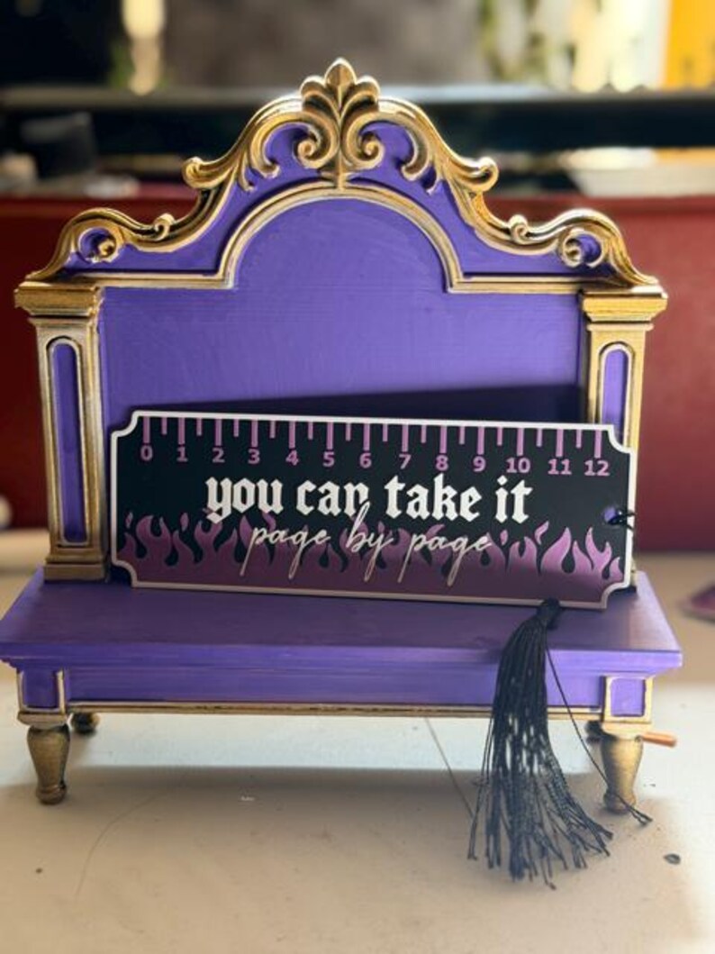May include: A purple and gold display stand featuring a rectangular bookmark with a black and purple flame pattern. The bookmark displays a 12-inch ruler along the top edge and the text "you can take it page by page" with a black tassel.
