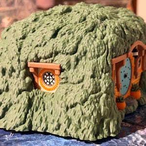 May include: A small, molded plastic structure resembling a hobbit-style earth house. The exterior is textured to look like green foliage or grass, featuring a round light blue door, a circular window with a grid pattern, and small orange planters.