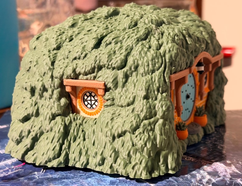May include: A small, molded plastic structure resembling a hobbit-style earth house. The exterior is textured to look like green foliage or grass, featuring a round light blue door, a circular window with a grid pattern, and small orange planters.