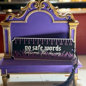 May include: A purple and gold display stand featuring a rectangular bookmark with a 12-inch ruler design. The bookmark is black with purple flames and white text that reads, "no safe words between the covens," and includes a black tassel.