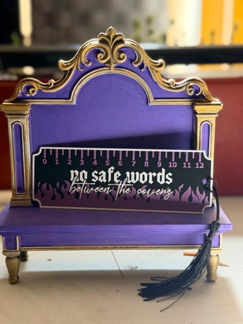 May include: A purple and gold display stand featuring a rectangular bookmark with a 12-inch ruler design. The bookmark is black with purple flames and white text that reads, "no safe words between the covens," and includes a black tassel.