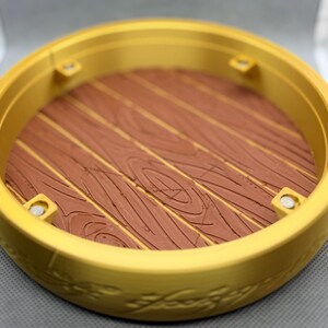 May include: A circular gold-colored display base featuring a raised rim with embossed script detailing. The interior floor of the base has a brown, textured surface mimicking the appearance of wooden planks, with four small silver-colored circular magnets.