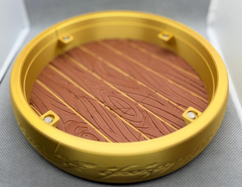 May include: A circular gold-colored display base featuring a raised rim with embossed script detailing. The interior floor of the base has a brown, textured surface mimicking the appearance of wooden planks, with four small silver-colored circular magnets.